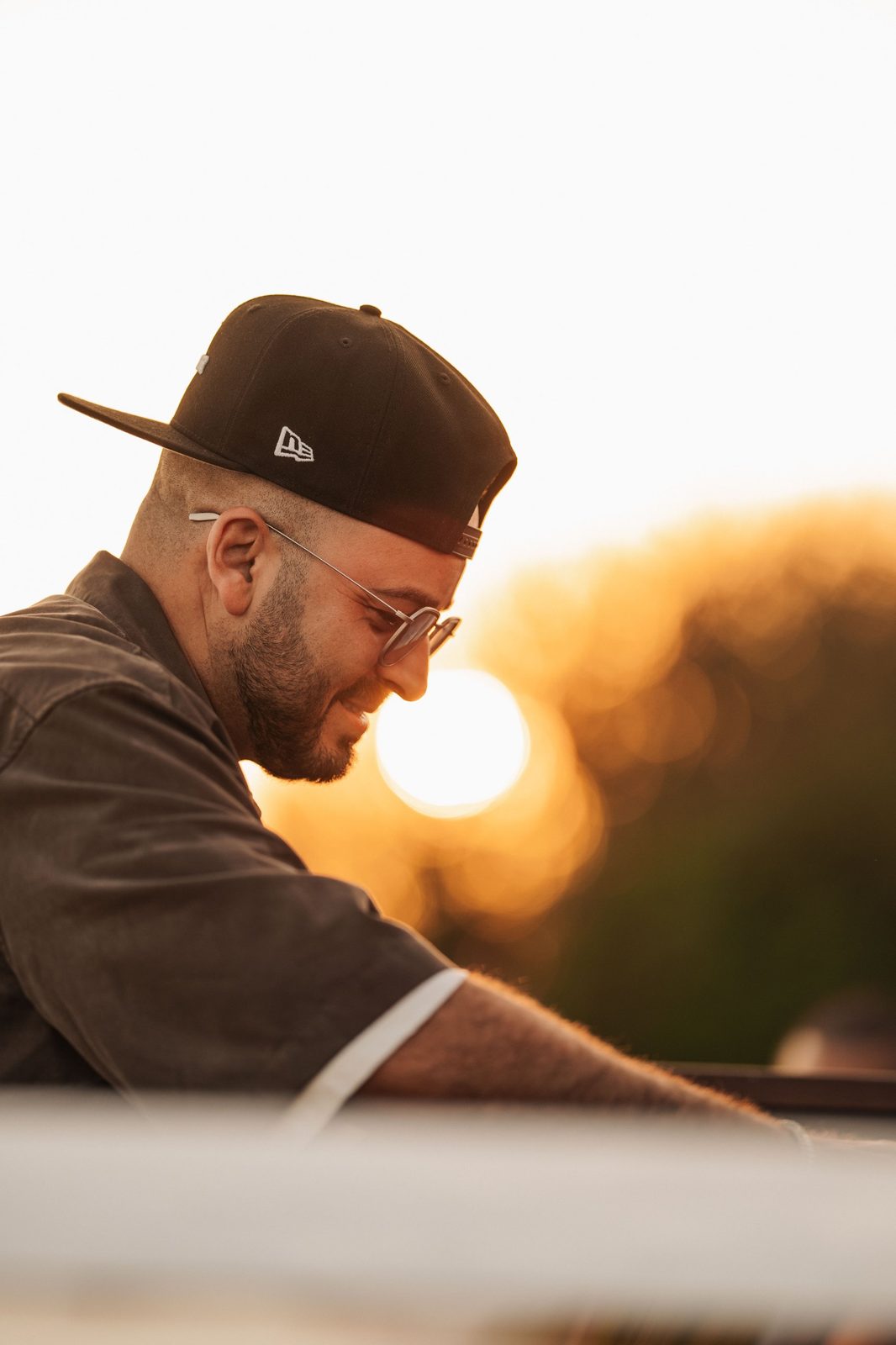 ERAN smiling at the decks at sunset, in a black cap and round glasses
